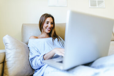 Porträt eines lächelnden jungen Mannes, der auf der Couch sitzt und einen Laptop benutzt - KIJF01483