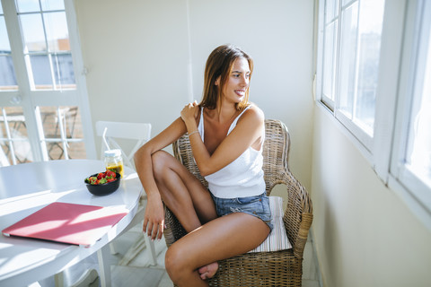 Junge Frau sitzt zu Hause auf einem Korbstuhl und schaut durch das Fenster, lizenzfreies Stockfoto