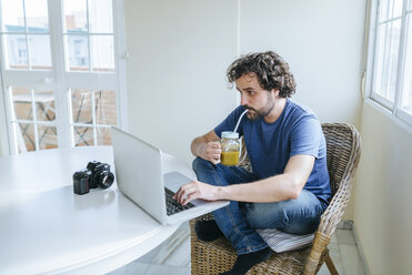 Mann trinkt Gemüsesaft, während er zu Hause einen Laptop benutzt - KIJF01496