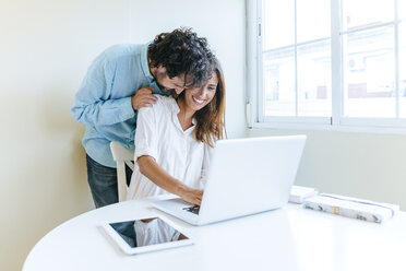 Mann umarmt Frau bei der Arbeit mit Laptop - KIJF01498