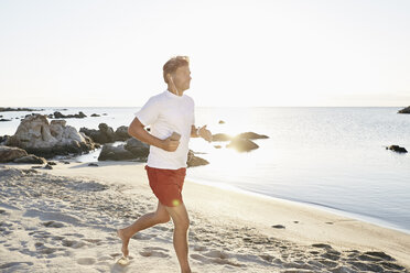 Älterer Mann joggt am Strand, während er mit Smartphone und Kopfhörern Musik hört - PDF01240