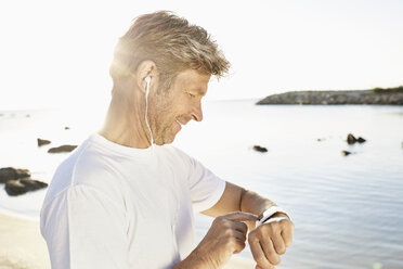 Älterer Mann mit Kopfhörer, der nach dem Joggen am Strand seine Smartwatch überprüft - PDF01241