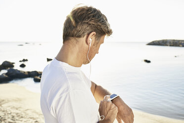 Älterer Mann mit Kopfhörer, der nach dem Joggen am Strand seine Smartwatch überprüft - PDF01242