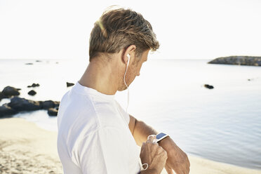 Mature man with earphone checking his smartwatch after yogging on the beach - PDF01242