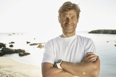 Porträt eines lächelnden reifen Mannes mit Smartwatch und Kopfhörern am Strand - PDF01243
