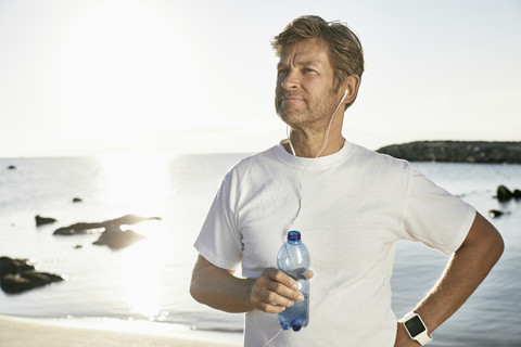 Porträt eines reifen Mannes mit Smartwatch und Kopfhörern, der nach dem Joggen am Strand Wasser trinkt, lizenzfreies Stockfoto