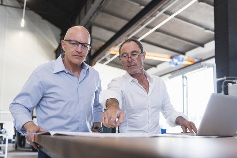 Zwei Geschäftsleute betrachten einen Plan auf einem Tisch in einer Fabrik, lizenzfreies Stockfoto