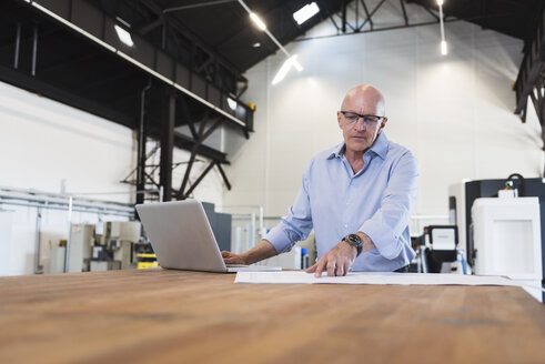 Mann mit Laptop betrachtet einen Plan auf einem Tisch in einer Fabrik - DIGF02495