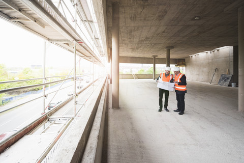 Zwei Männer mit Plan, die Sicherheitswesten tragen, unterhalten sich in einem im Bau befindlichen Gebäude, lizenzfreies Stockfoto