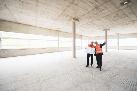 Zwei Männer mit Plan, die Sicherheitswesten tragen, unterhalten sich in einem im Bau befindlichen Gebäude, lizenzfreies Stockfoto