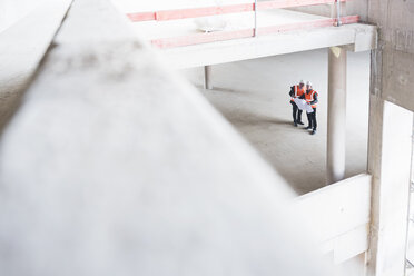 Two men with plan wearing safety vests talking in building under construction - DIGF02530