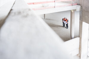Zwei Männer mit Plan, die Sicherheitswesten tragen, unterhalten sich in einem im Bau befindlichen Gebäude - DIGF02530