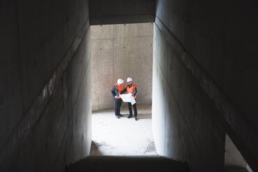 Two men with plan wearing safety vests talking in building under construction - DIGF02531