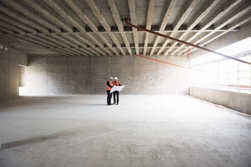 Two men with plan wearing safety vests talking in building under construction - DIGF02536