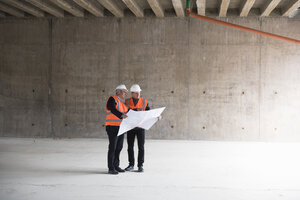 Zwei Männer mit Plan, die Sicherheitswesten tragen, unterhalten sich in einem im Bau befindlichen Gebäude - DIGF02537