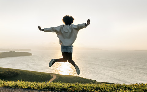 Junge Frau springt bei Sonnenuntergang an der Küste, lizenzfreies Stockfoto