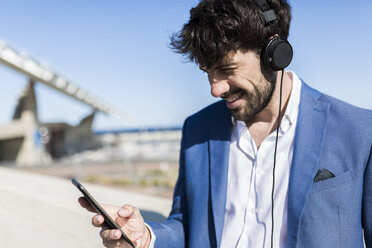 Young businessman wearing headphones looking at smartphone outdoors - GIOF02602