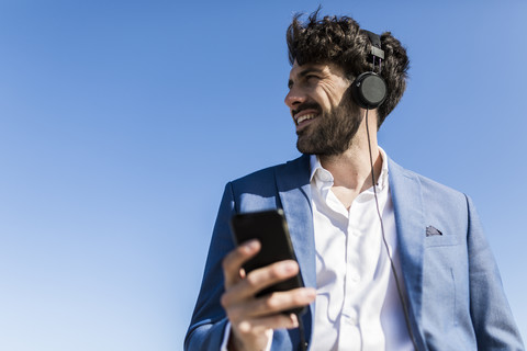 Junger Geschäftsmann mit Smartphone und Kopfhörern unter blauem Himmel, lizenzfreies Stockfoto