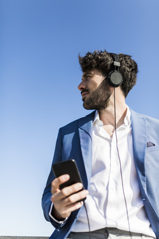 Junger Geschäftsmann mit Smartphone und Kopfhörern unter blauem Himmel, lizenzfreies Stockfoto