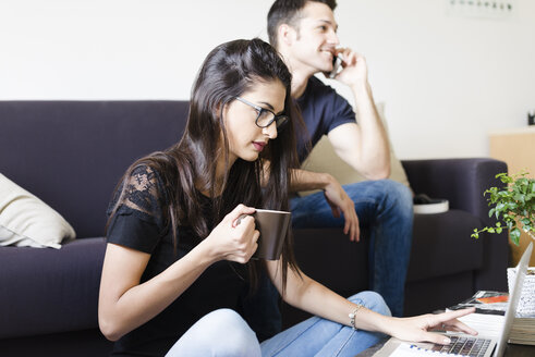 Junge Frau mit einer Tasse Kaffee am Laptop im Wohnzimmer, während ihr Freund telefoniert - FMOF00275