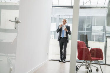 Confident businessman standing in office, eating take out food - FMKF04097