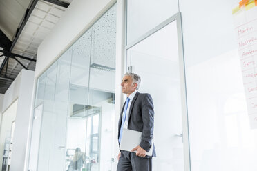 Mature businessman standing in office, holding laptop - FMKF04138