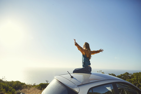 Südafrika, Kapstadt, Signal Hill, junge Frau sitzt auf dem Dach eines Autos und genießt den Blick auf das Meer, lizenzfreies Stockfoto