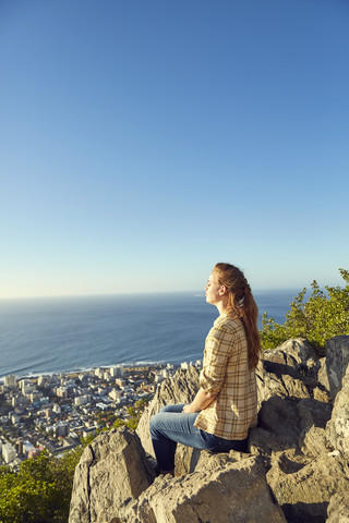 Südafrika, Kapstadt, Signal Hill, junge Frau sitzt auf einem Felsen mit Blick auf die Stadt und das Meer, lizenzfreies Stockfoto