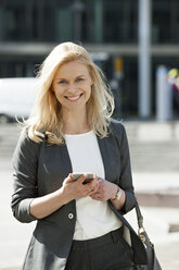 Porträt einer lächelnden blonden Geschäftsfrau mit Mobiltelefon und Handtasche - CHAF01877