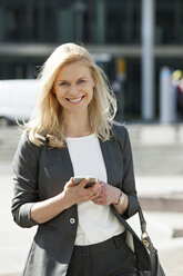 Portrait of smiling blond businesswoman with cell phone and handbag - CHAF01877