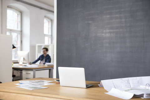 Desk in office with laptop, photographies and blueprint and man in backgound stock photo