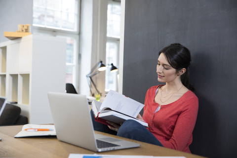 Woman with book and laptop at desk in office stock photo