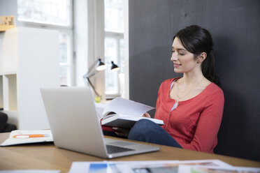 Woman with book and laptop at desk in office - FKF02285