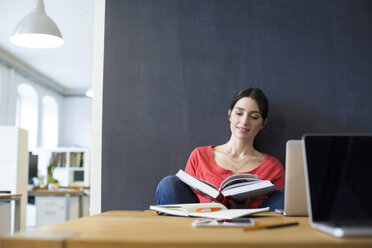 Woman with book and laptop at desk in office - FKF02325