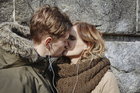 Junges Paar teilt sich Ohrstöpsel und küsst sich an einer Steinmauer - ANHF00033