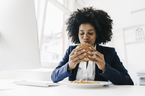 Young businesswoman eating hamburger at her desk - KNSF01388