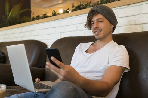 Junger Mann benutzt Handy und Laptop in einem Cafe - KKAF00823