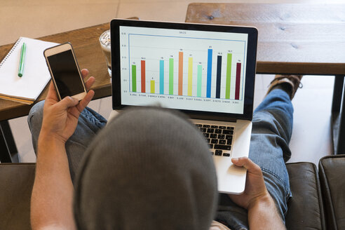 Young man using cell phone and laptop with bar chart in a cafe - KKAF00824