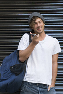 Smiling young man using cell phone at roller shutter - KKAF00850