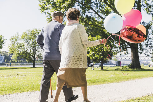 Rückansicht eines älteren Paares mit Luftballons, das in einem Park spazieren geht - UUF10645