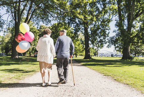 Back view of senior couple with balloons strolling in a park - UUF10646