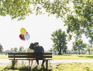 Happy senior couple with balloons sitting on bench in a park - UUF10648