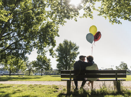 Glückliches älteres Paar mit Luftballons auf einer Bank in einem Park sitzend - UUF10649