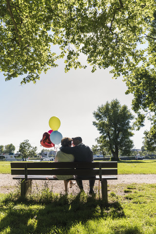 Glückliches älteres Paar mit Luftballons, die sich auf einer Bank in einem Park küssen, lizenzfreies Stockfoto
