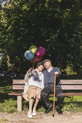 Glückliches älteres Paar mit Luftballons auf einer Bank in einem Park sitzend - UUF10651