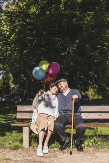 Happy senior couple with balloons sitting on bench in a park - UUF10651