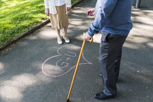 Senior couple drawing love heart with initials on tarmac - UUF10655