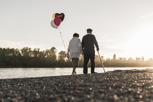 Rückansicht eines älteren Paares mit Luftballons, das am Abend am Flussufer spazieren geht - UUF10685