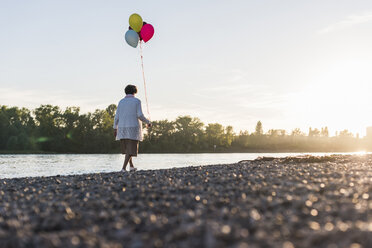 Rückenansicht einer älteren Frau mit Luftballons, die am Flussufer spazieren geht - UUF10686