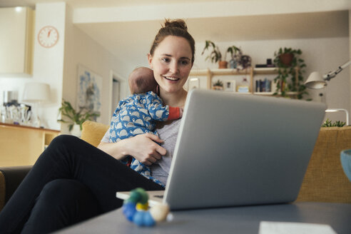 Mother using laptop and holding her newborn baby at home - MFF03581